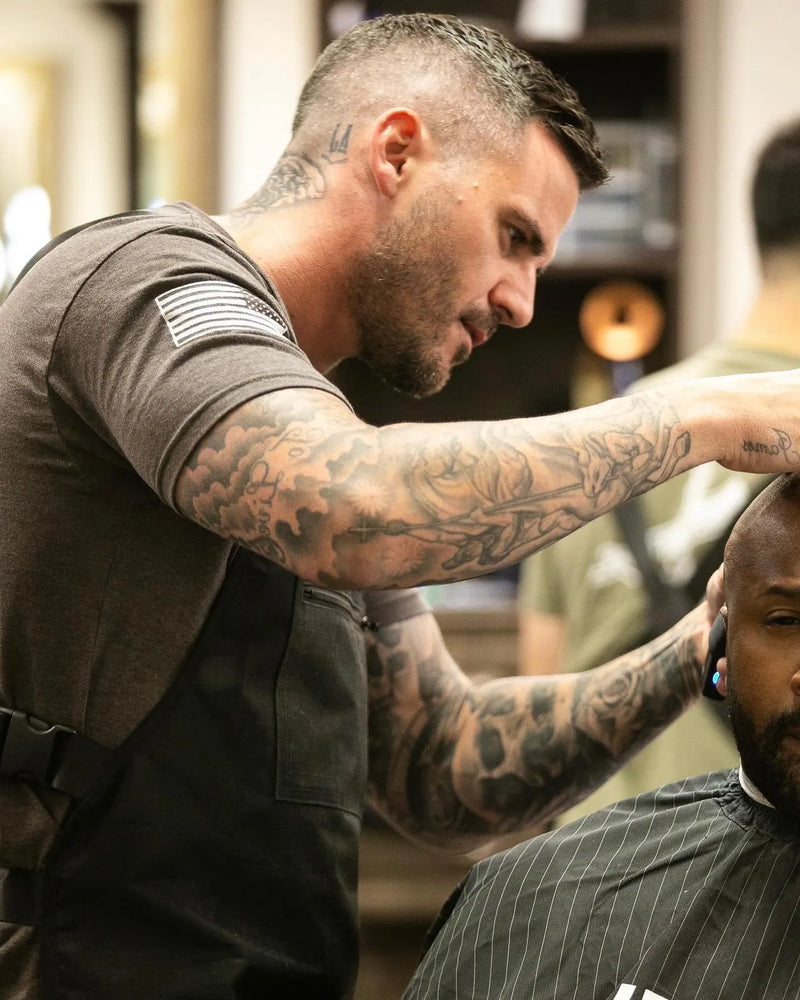 Man with tattoos cutting another man's hair in a barbershop.